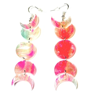 May include: A pair of dangle earrings featuring a moon phase design. Each earring has a silver hook and is composed of a series of colorful, circular and crescent-shaped elements in shades of pink, orange, and white with glitter accents.