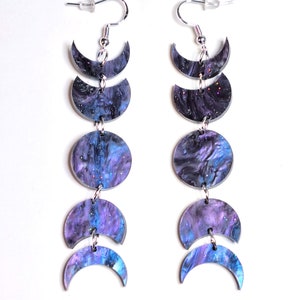 Può includere: Un paio di orecchini pendenti con un design ispirato alla galassia. Ogni orecchino presenta cinque forme di luna crescente in tonalità di viola, blu e nero con accenti di glitter argentati. Le lune sono collegate da catene argentate.