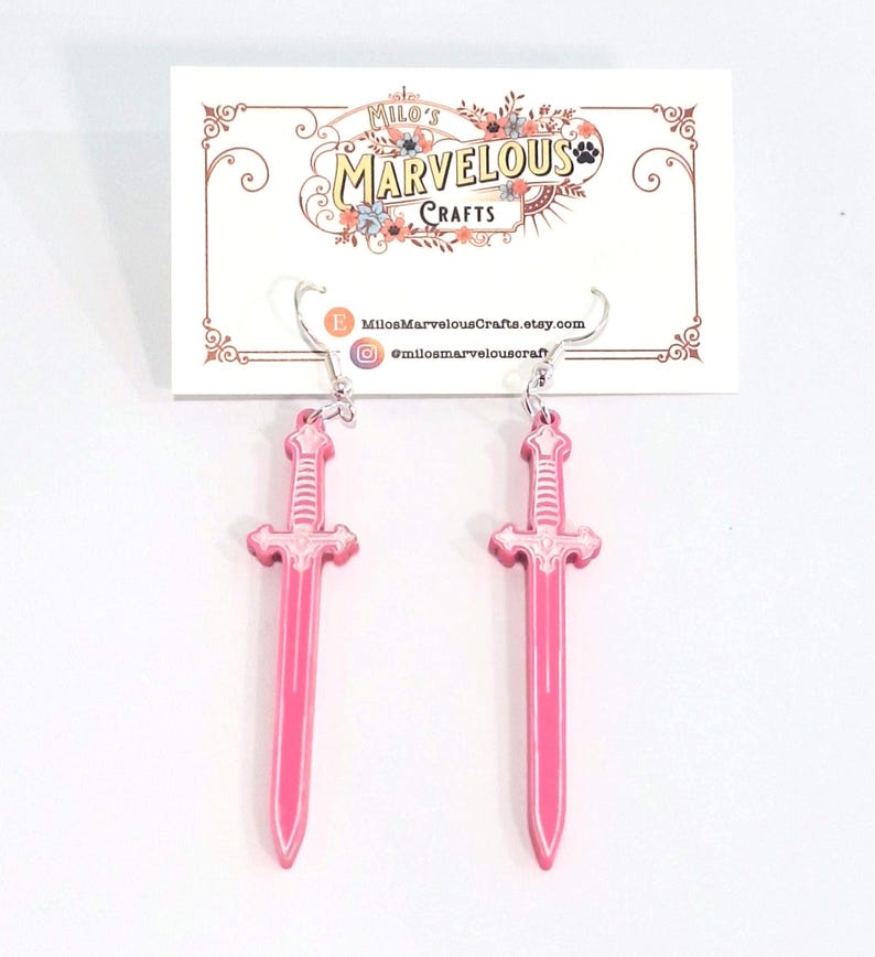 Op de afbeelding: Een paar roze zwaardvormige oorbellen met zilverkleurige oorhaken. De oorbellen worden getoond tegen een witte achtergrond met een kaart met de tekst "Milo's Marvelous Crafts".