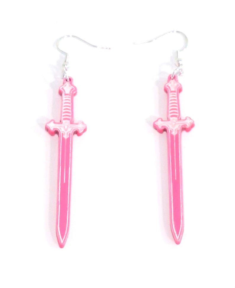 Op de afbeelding: Een paar roze zwaardvormige oorbellen met zilverkleurige haken. De oorbellen hebben een gedetailleerd ontwerp met een witte omtrek, en de zwaarden zijn felroze. De oorbellen worden tegen een witte achtergrond getoond.