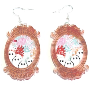 Op de afbeelding: Een paar oorbellen met een ovaal frame-ontwerp in koperkleur. De frames bevatten een bloemenpatroon en witte spookachtige figuren. De oorbellen hebben zilverkleurige haken.