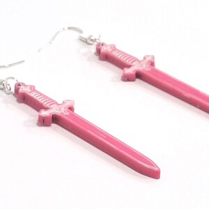Op de afbeelding: Een paar roze dolkvormige oorbellen met zilverkleurige oorhaken. De oorbellen hebben een levendige roze tint en een gedetailleerd handvatontwerp. De oorbellen worden op een witte achtergrond gepresenteerd.