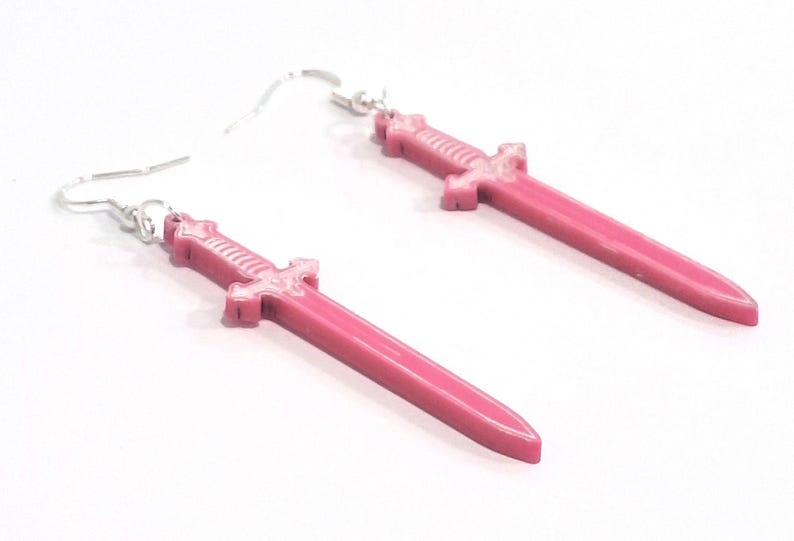 Op de afbeelding: Een paar roze dolkvormige oorbellen met zilverkleurige oorhaken. De oorbellen hebben een levendige roze tint en een gedetailleerd handvatontwerp. De oorbellen worden op een witte achtergrond gepresenteerd.