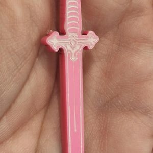 Op de afbeelding: Een roze zwaardvormige oorbel met witte details. De oorbel is een miniatuur replica van een zwaard, met een pareerstang en een gedetailleerde greep. De oorbel is opgehangen aan een zilverkleurige oorhaak.