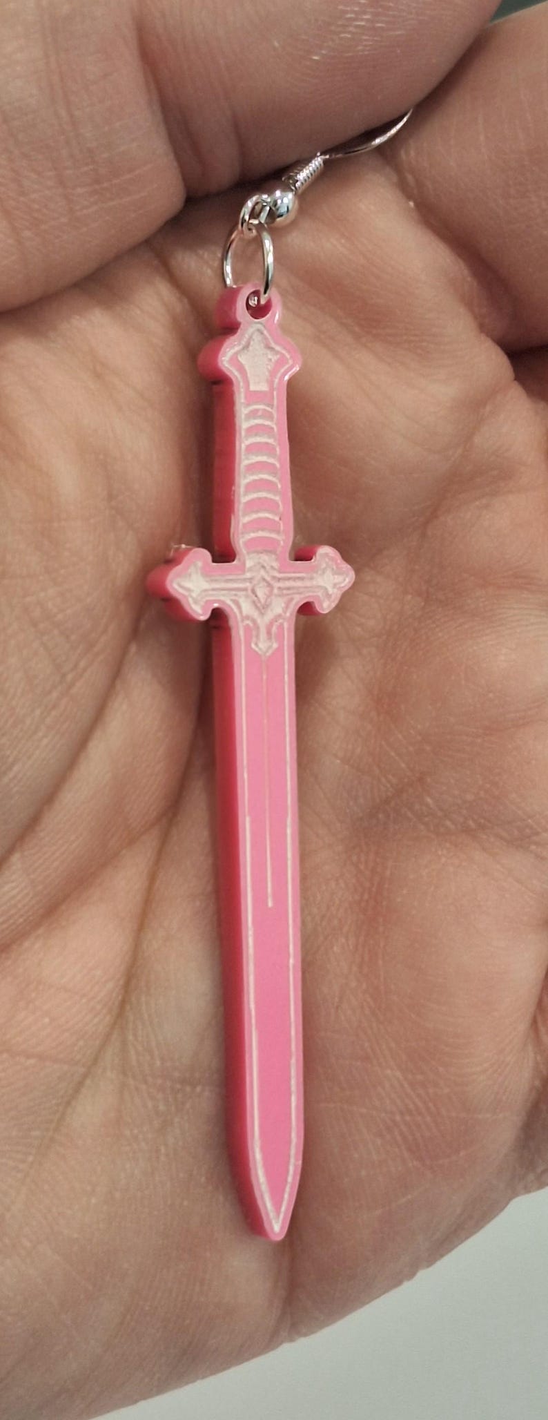 Op de afbeelding: Een roze zwaardvormige oorbel met witte details. De oorbel is een miniatuur replica van een zwaard, met een pareerstang en een gedetailleerde greep. De oorbel is opgehangen aan een zilverkleurige oorhaak.