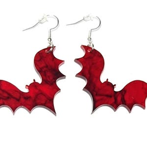May include: A pair of red bat-shaped earrings with a marbled effect. The earrings are made of acrylic and have silver hooks.