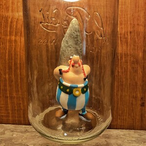 Peut inclure: Lampe en bocal en verre transparent avec un raccord métallique et une ampoule à l'intérieur. La lampe contient une figurine d'un personnage de dessin animé. La lampe est fixée sur une surface en bois. Le texte "mambo na mambo Tarros de Luz mambo na mambo.com" est visible.