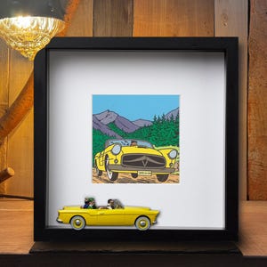 Puede incluir: Una impresi&oacute;n enmarcada de un coche descapotable amarillo con un fondo de monta&ntilde;a. Un coche amarillo en miniatura con dos figuras se encuentra frente a la impresi&oacute;n enmarcada. El marco es negro y el fondo es de madera.