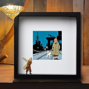 Könnte beinhalten: Gerahmter Druck mit einer Comic-Illustration einer Person in einem beigefarbenen Mantel und einem weißen Hund auf Bahngleisen. Im Hintergrund ist eine Hafenszene zu sehen. Der Rahmen ist schwarz. Der Druck wird auf einer Holzoberfläche präsentiert.