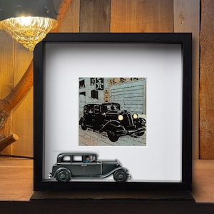May include: A framed artwork featuring a black vintage car illustration with Japanese text. A miniature gray car model sits in front of the frame. The artwork is displayed on a wooden surface, illuminated by a lamp.