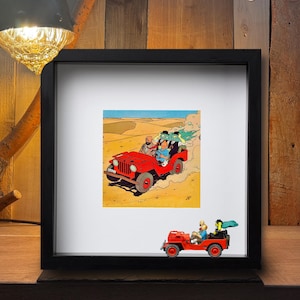 Könnte beinhalten: Gerahmtes Kunstwerk mit einem roten Jeep und Comicfiguren in einer Wüstenlandschaft. Ein Spielzeug-Jeep befindet sich vor dem Rahmen. Der Hintergrund ist eine Holzoberfläche.
