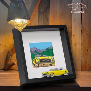 Puede incluir: Un marco negro con una impresi&oacute;n de un coche descapotable amarillo frente a un paisaje monta&ntilde;oso. Un peque&ntilde;o coche amarillo est&aacute; fijado al marco. El conjunto est&aacute; sobre una superficie de madera.