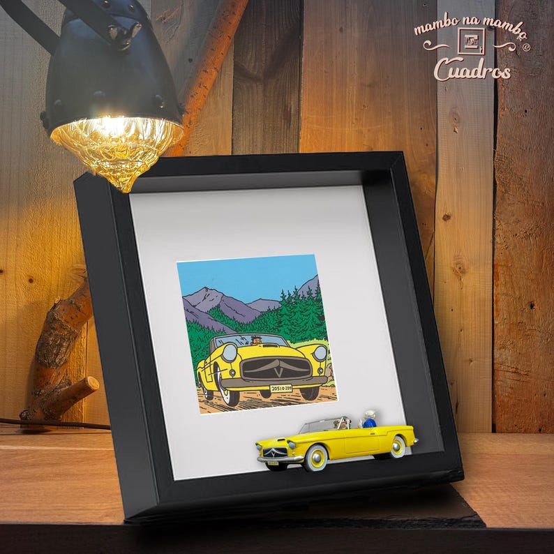 Puede incluir: Un marco negro con una impresi&oacute;n de un coche descapotable amarillo frente a un paisaje monta&ntilde;oso. Un peque&ntilde;o coche amarillo est&aacute; fijado al marco. El conjunto est&aacute; sobre una superficie de madera.