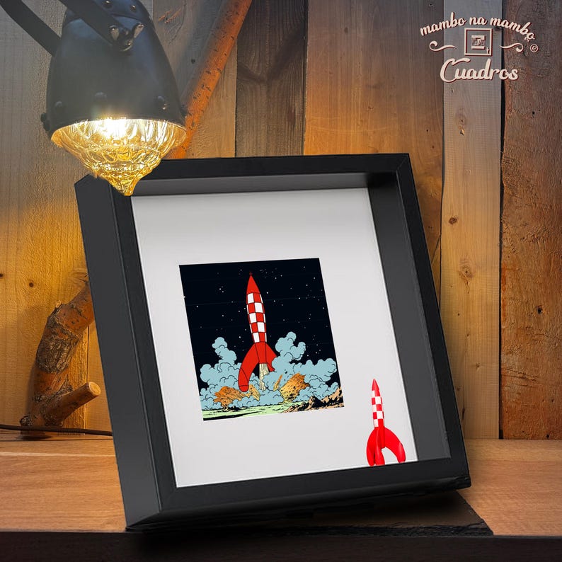 May include: A square black shadow box frame containing a white mount and a print of a red and white chequered rocket ship launching into a dark starry sky, with a smaller matching rocket graphic positioned in the bottom right corner of the mount.