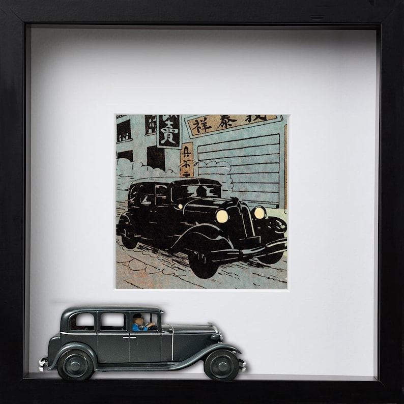 May include: A framed artwork featuring a black vintage car illustration in a Japanese style. Below the artwork is a miniature model of a black car with a driver. The artwork is in a black frame.