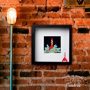 May include: A square black shadow box frame containing a print of a red and white chequered rocket ship launching into a dark starry sky with light blue smoke clouds. A small, matching red and white rocket ship figurine sits inside the frame.