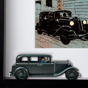 May include: A framed image featuring a vintage black car model and a comic strip illustration of a similar car. The model car is dark green with a driver figure. The comic strip shows a black car on a snowy street.