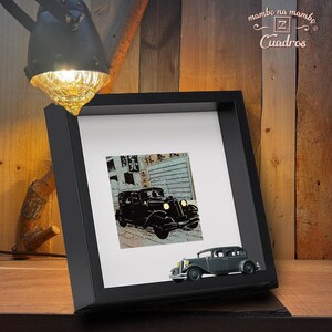 May include: A black framed print featuring a vintage car illustration, with a miniature car model attached to the frame. The print has Japanese text and is set against a wooden background. The frame is angled, with a lamp in the upper left corner.