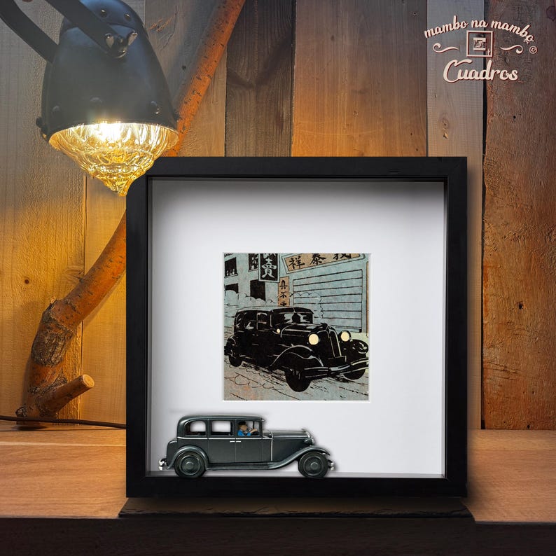 May include: A framed artwork featuring a black vintage car illustration with Japanese text. A miniature gray car model sits in front of the frame. The artwork is displayed on a wooden surface, illuminated by a lamp.