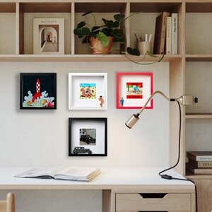 May include: A collection of framed art prints displayed on a white desk. The prints feature various illustrations, including a rocket launch, beach scene, cyclist, and vintage car. The frames are black, white, and red.