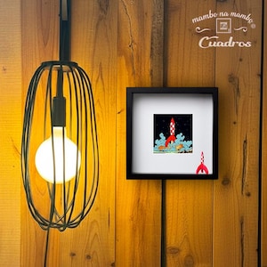 May include: A black wire cage pendant light with a glowing round bulb next to a square black frame containing a print of a red and white rocket ship launching into space, with a small red rocket figurine placed in the corner of the frame.