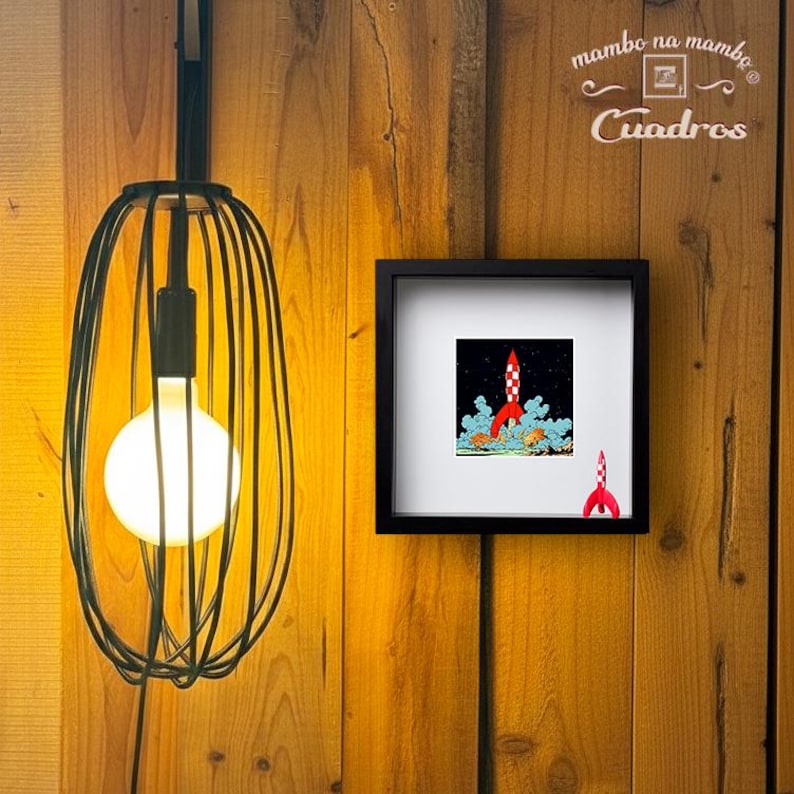 May include: A black wire cage pendant light with a glowing round bulb next to a square black frame containing a print of a red and white rocket ship launching into space, with a small red rocket figurine placed in the corner of the frame.
