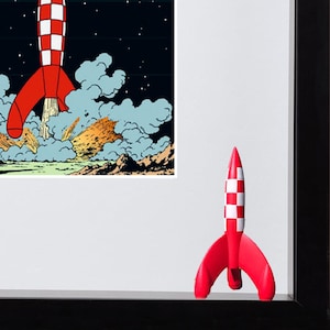 May include: A framed print featuring a red and white checkered rocket ship launching into a dark sky with clouds of smoke, accompanied by a small, three-dimensional red and white checkered rocket figurine standing on the black frame edge.