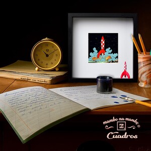 May include: A framed square print featuring a red and white rocket ship launching into a dark starry sky, displayed on a desk with a yellow analogue alarm clock, a notebook with handwritten text, an inkwell, a pen, and a small rocket figurine.