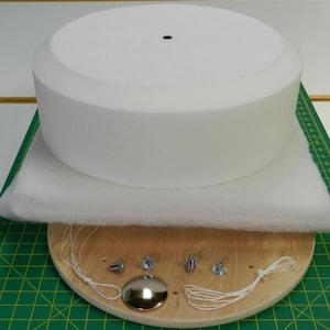 May include: A white foam cake dummy with a round wooden base, white string, and metal hardware. The cake dummy is on top of a white batting pad.