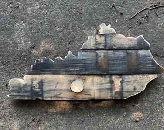 May include: A wooden outline of the state of Kentucky, made from reclaimed bourbon barrel staves. The outline is dark brown and black with a light brown circle in the center.
