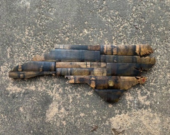 May include: A wooden map of North Carolina, made from reclaimed bourbon barrel staves. The map is a dark brown color with some lighter brown and black accents. The map is a unique and rustic piece of home decor.