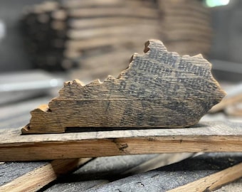 May include: A wooden cutout of the state of Kentucky, shaped like the state outline. The wood is dark brown and has a rustic, weathered look. The cutout is sitting on a piece of wood that appears to be a barrel stave.