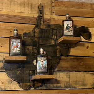 May include: A wooden shelf shaped like the state of West Virginia, with three shelves. Each shelf holds a bottle of liquor with a unique label. The bottles are all brown glass and have a vintage look.