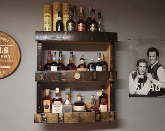 May include: A wooden wall-mounted shelf with three tiers, each holding various bottles of liquor. The shelf is made from reclaimed wood and has a rustic appearance. The bottles are arranged in a staggered pattern, with some bottles facing forward and others facing backward. The shelf is mounted on a gray wall.