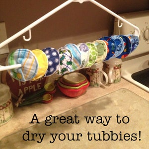 May include: A white plastic hanger with multiple colourful fabric circles hanging from it. The circles are made of soft fabric and have different patterns, including stripes, polka dots, and animals. The text "A great way to dry your tubbies!" is written below the hanger.