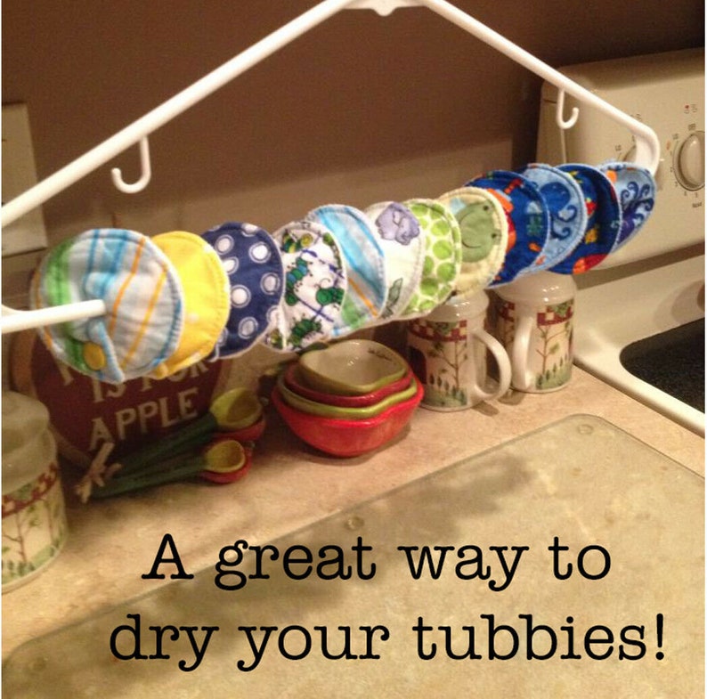 May include: A white plastic hanger with multiple colourful fabric circles hanging from it. The circles are made of soft fabric and have different patterns, including stripes, polka dots, and animals. The text "A great way to dry your tubbies!" is written below the hanger.