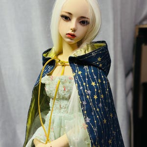 May include: A doll with long, white hair and a pale complexion, wearing a light green floral dress and a navy blue cape. The cape is adorned with gold stars and a yellow lining. The doll's cape is secured with a yellow cord.