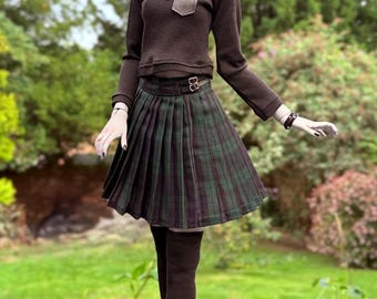 May include: A doll wearing a brown beret, long-sleeved shirt with a pocket, and a pleated plaid skirt. The doll is also wearing black tights, a necklace, and black and white sneakers. The doll is standing on a gray surface with a green background.