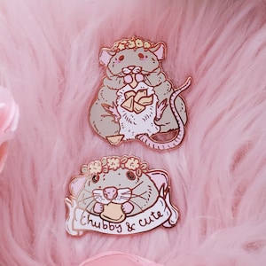 May include: Two enamel pins featuring a cartoon rat wearing a flower crown. The first pin shows the rat holding a piece of cheese. The second pin shows the rat holding a piece of cheese and a banner that says "chubby & cute".