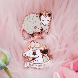 May include: Two enamel pins featuring a possum. One pin is a white possum with a flower crown and the text "Scream all day". The other pin is a green possum with a flower crown.