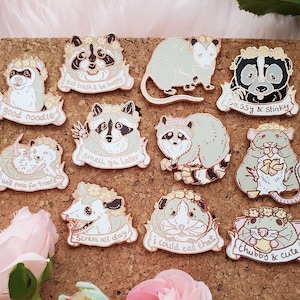 May include: A corkboard with 11 enamel pins featuring various animals, including raccoons, opossums, and rats. Each pin has a different saying, such as "good noodle", "smell ya later", and "chubby & cute".