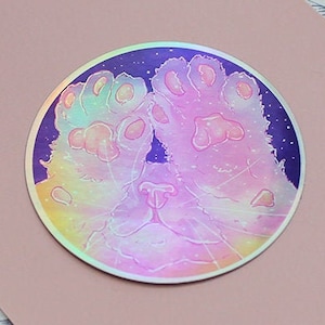 May include: A round iridescent sticker with a cat's paws and face. The sticker has a holographic finish and a galaxy-like background.