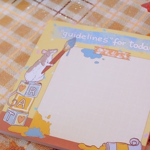 May include: A square notepad with a light yellow background and the text "guidelines for today". The notepad features a cartoon rat with a paintbrush, paint tubes, and the letters R, A, and T on blocks. The notepad is surrounded by autumn leaves and acorns.