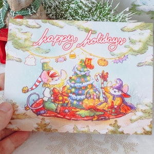 May include: A Christmas card with a blue background featuring three mice on a red sled. The mice are pulling a decorated Christmas tree. The card says "Happy Holidays" in pink lettering.
