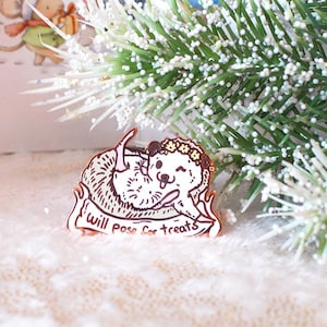 May include: A wooden pin with a cartoon illustration of a dog wearing a flower crown and a banner that reads "Will pass out treats". The pin is on a white background with a sprig of green pine.
