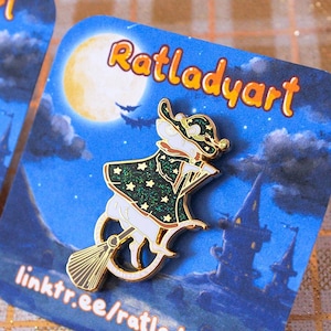 May include: A gold-colored enamel pin featuring a white mouse wearing a green and gold witch's hat and robe, riding a broomstick. The pin is set against a blue background with a full moon, stars, and a castle in the distance. The text "Ratladyart" is printed on the card.