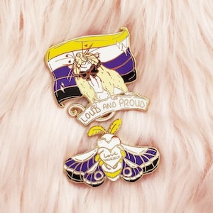 May include: Gold enamel pin featuring a purple, yellow, and white flag with a rat wearing a bow tie and the text "Loud and Proud". Below the flag is a gold moth with a heart that says "Love Yourself".