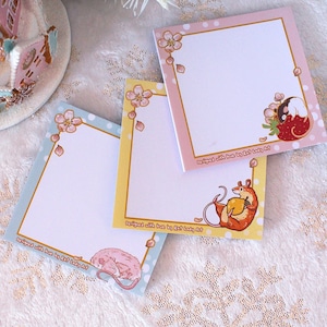 May include: Three pastel colored square sticky note pads with a white background and a decorative border. The border features a floral design with pink flowers and gold dots. Each pad has a different illustration of a mouse. One mouse is holding a strawberry, another is holding a piece of cheese, and the third is sleeping. The text on the bottom of each pad reads "Everything with you is so much better, my little kitty cat."