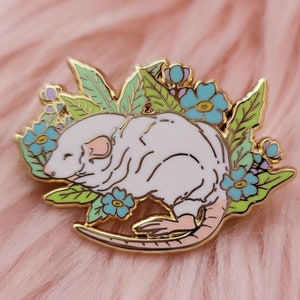 May include: A gold-tone enamel pin featuring a white rat with pink ears and tail, surrounded by green leaves and blue flowers.