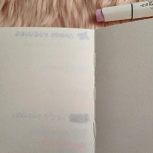 May include: Open blank notebook with stitched binding, pages are white. A white marker with a lavender cap rests on the top right corner. The background is a soft, pink, fuzzy material. Faint text is visible on the notebook pages.
